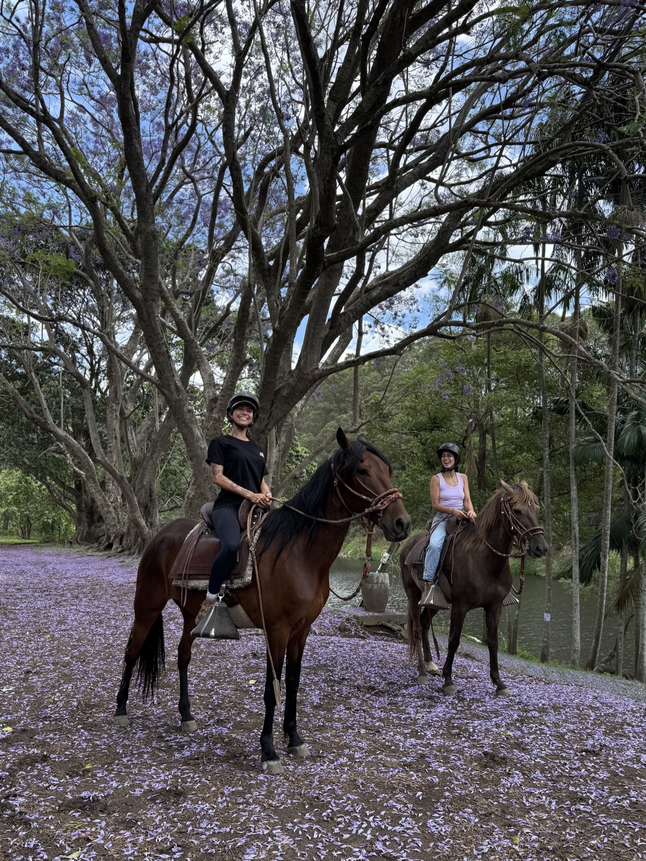 Two smiling riders on horseback beneath the jacarandas, purple petals on the ground at Historic Rivermill